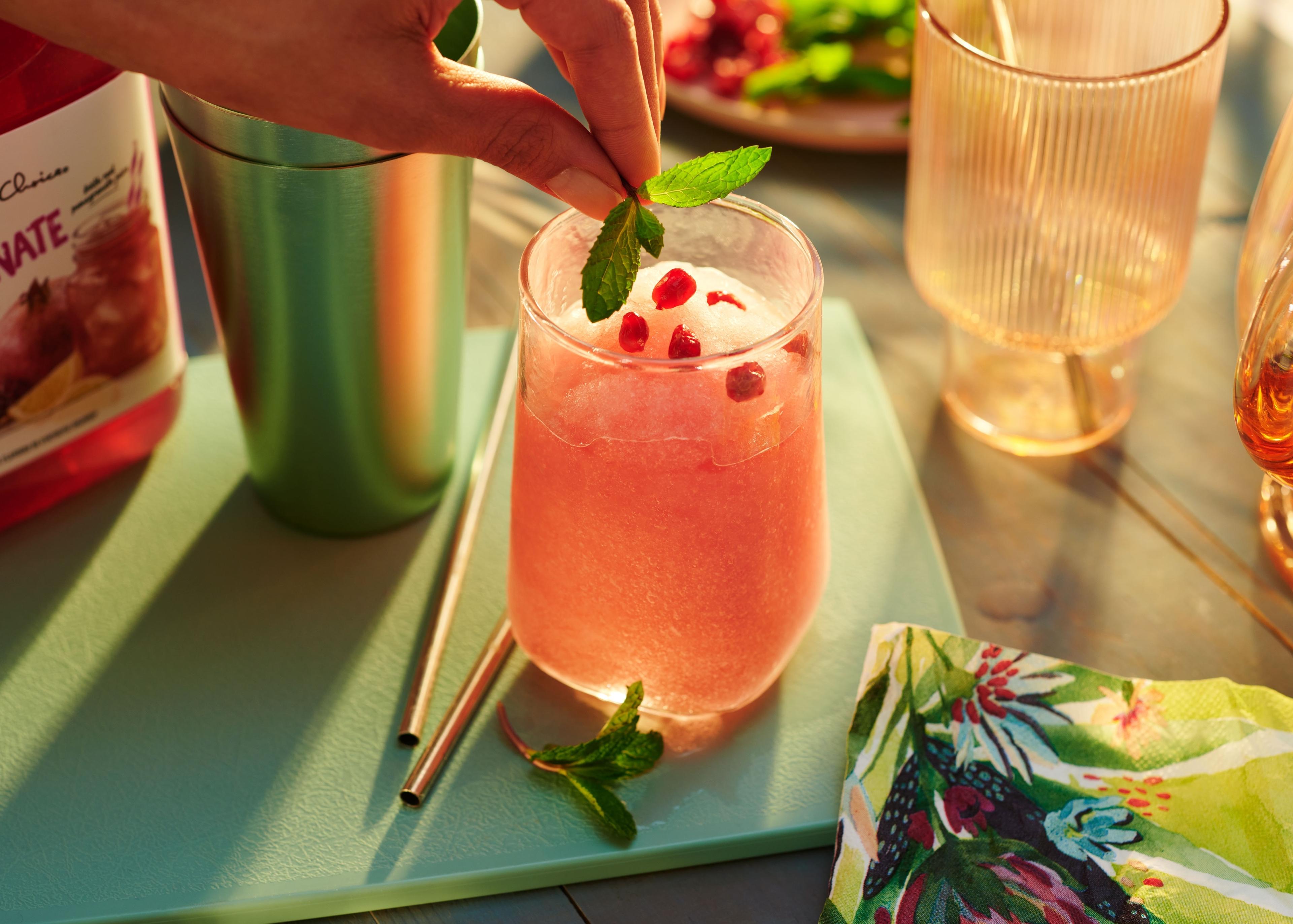 beautifully styled cranberry and crushed ice drink with hand gently placing a final touch of mint leaves. The glass is sitting on a morning-lit counter-top with cutting board and straws, napkins.