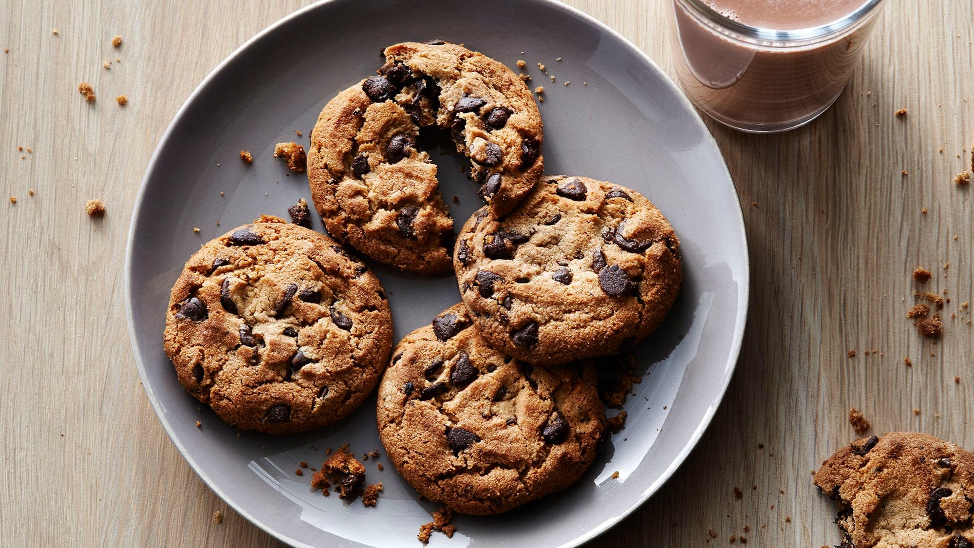 Artfully photographed “the Decadent” PC chocolate chip cookies on a plate. One of them is temptingly broken in half, with crumbs scattered on the plate.