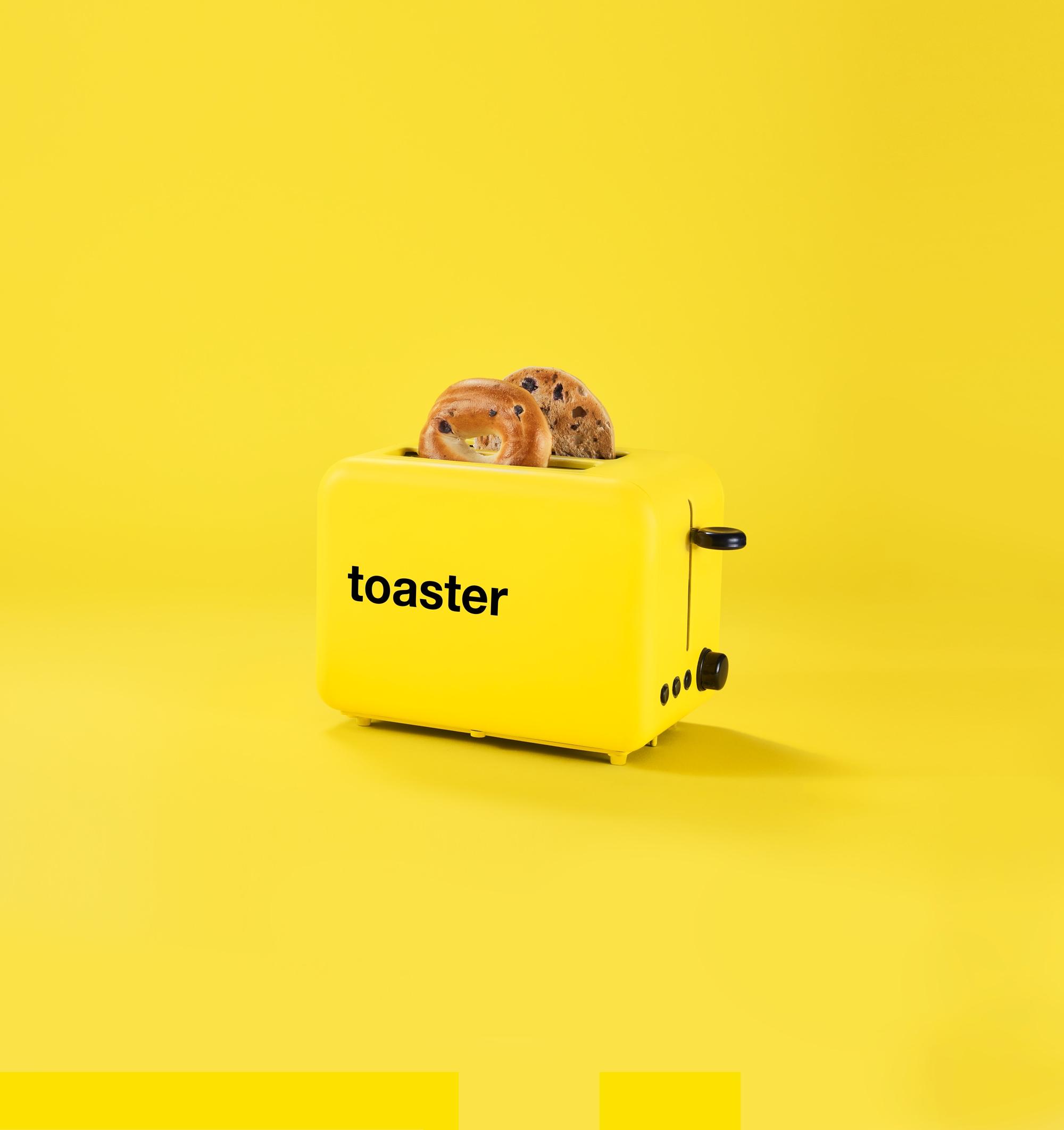 A yellow background with a yellow toaster. There are two blueberry bagels popped up out of the toaster. The word “toaster” is printed on the toaster in true no name style.